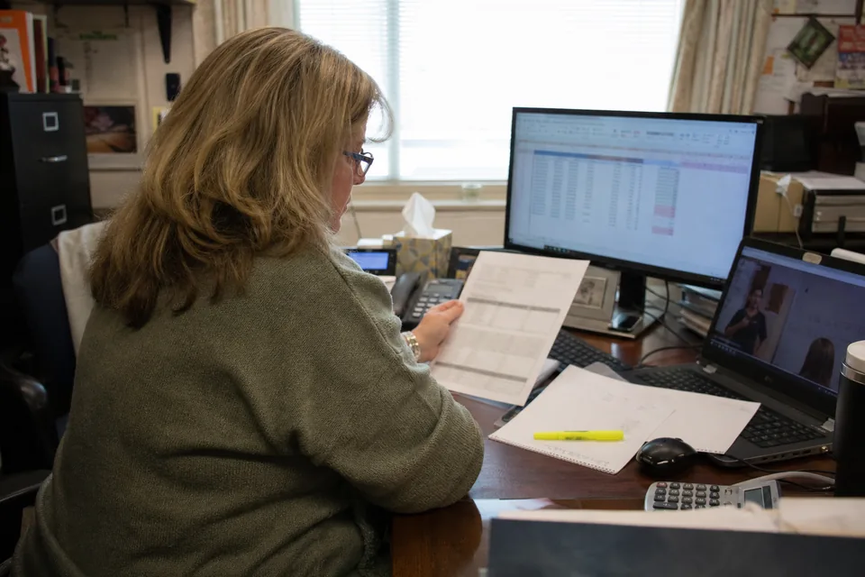 District technology coordinator comparing a printed software renewal quote with a spreadsheet, a video lesson paused on a laptop beside it