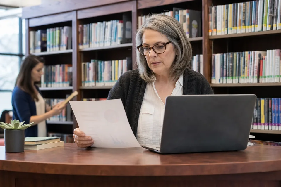 School librarian at the circulation desk comparing a printed renewal quote with a laptop