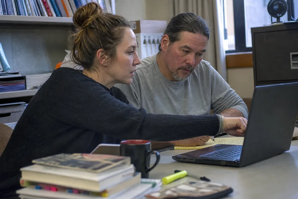 Two literacy teachers at a workroom table reviewing a laptop report next to tabbed paperback novels and a legal pad