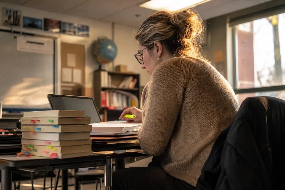Reading teacher in an empty classroom reviewing written answers on a laptop beside a stack of novels